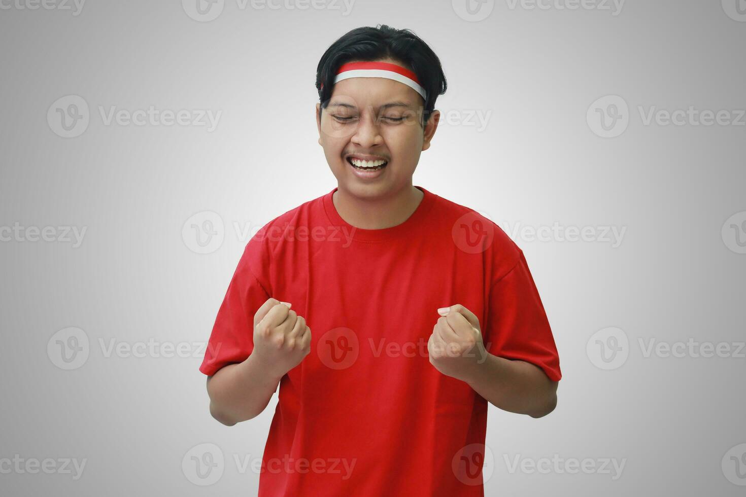 Portrait of attractive Asian man in t-shirt with red and white ribbon on head, raising his fist, celebrating Indonesia's independence day. Isolated image on gray background photo