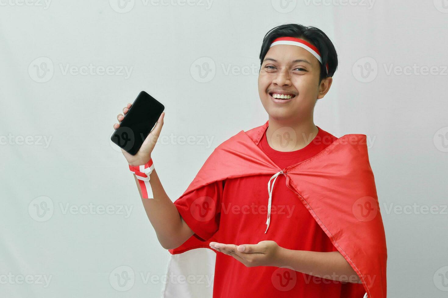 Portrait of attractive Asian man in t-shirt with red white ribbon on head with flag on his shoulder as a cloak, presenting a blank screen mobile phone. Isolated image on gray background photo