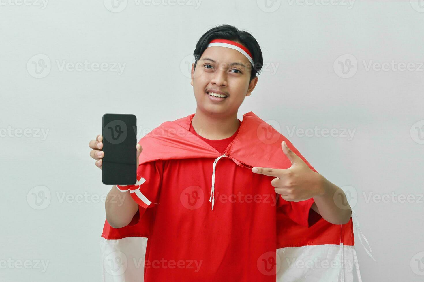 Portrait of attractive Asian man in t-shirt with red white ribbon on head with flag on his shoulder as a cloak, presenting a blank screen mobile phone. Isolated image on gray background photo