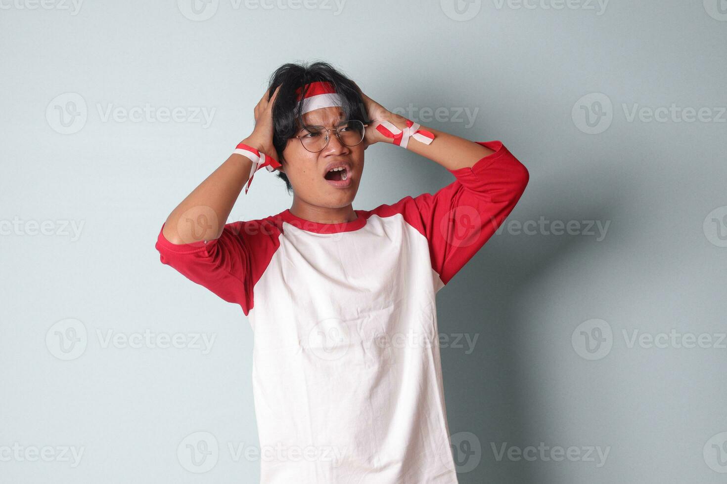 retrato de atractivo asiático hombre en camiseta con rojo y blanco cinta en cabeza, teniendo un migraña, conmovedor su templo. dolor de cabeza concepto. aislado imagen en gris antecedentes foto