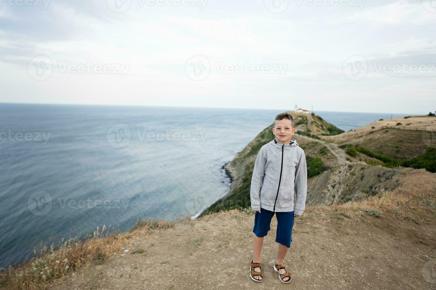 Little boy standing on top of a mountain and looking camera against lighthouse in sea. Cape Emine, Black sea coast, Bulgaria. photo