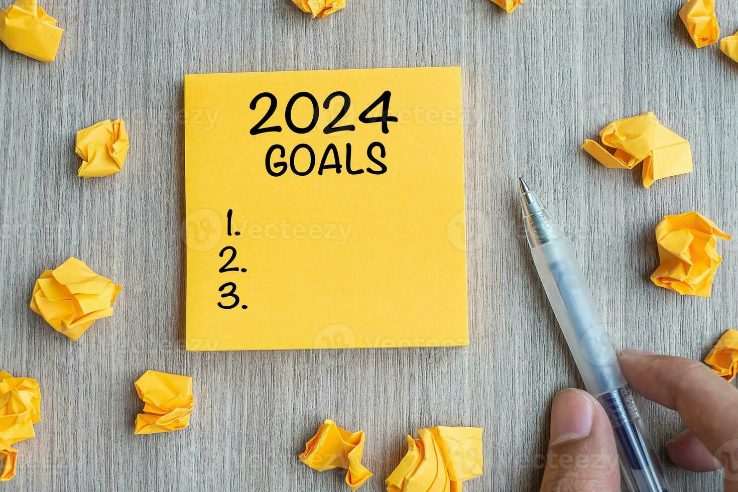 2024 Goal word on yellow note with Businessman holding pen and crumbled paper on wooden table background. New Year New Start, Resolutions, Strategy, mission concept photo