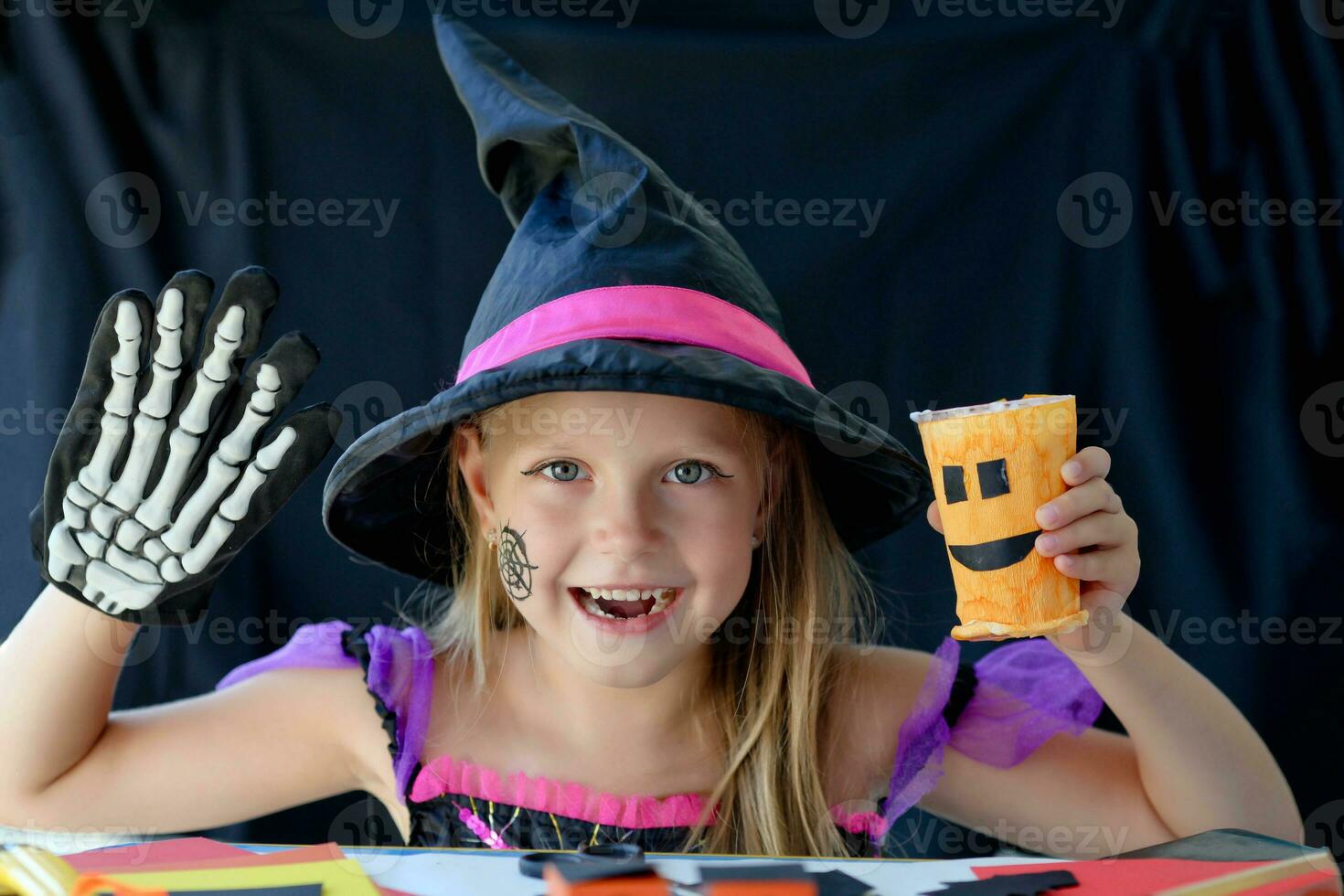 A little girl in a witch costume looks at the camera and laughs, holding a glass pumpkin and a skeleton glove in her hands. Halloween holiday, crafts, handmade. photo