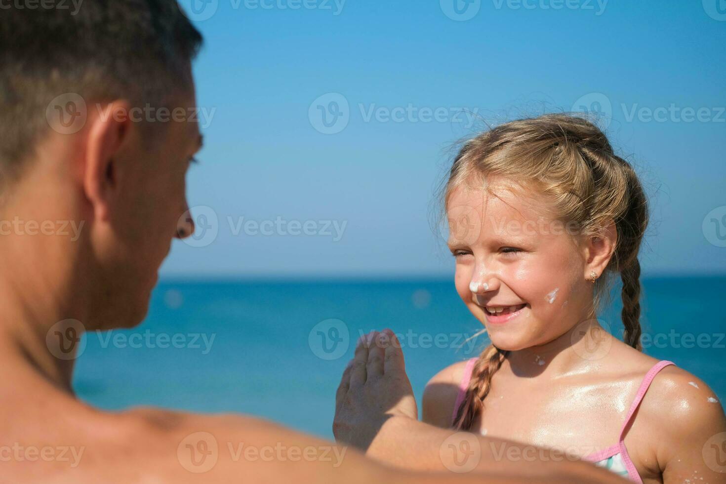 padre aplicando protector crema a su hija cara a el playa. hombre mano participación protector solar loción en bebé rostro. linda pequeño niña con un Dom bloquear por el mar . Copiar espacio. foto
