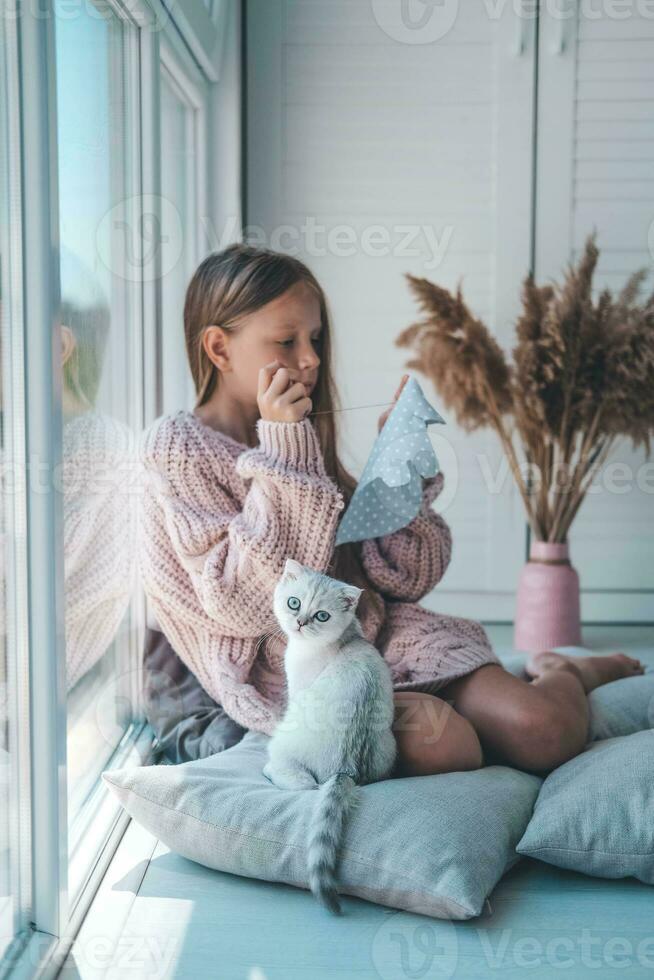 The child sews a soft toy near the window and plays with the kitten. The girl sews up a soft toy of an elephant. Handmade, hobby. photo