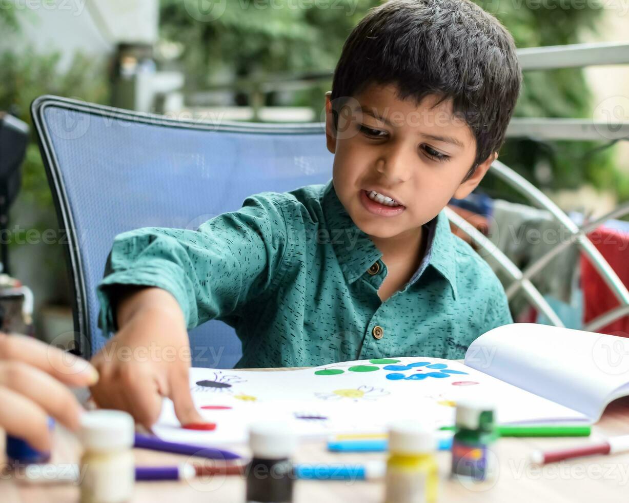 inteligente indio pequeño chico realizar pulgar pintura con diferente vistoso agua color equipo durante el verano vacaciones, linda indio niño haciendo vistoso pulgar pintura dibujo en de madera mesa foto