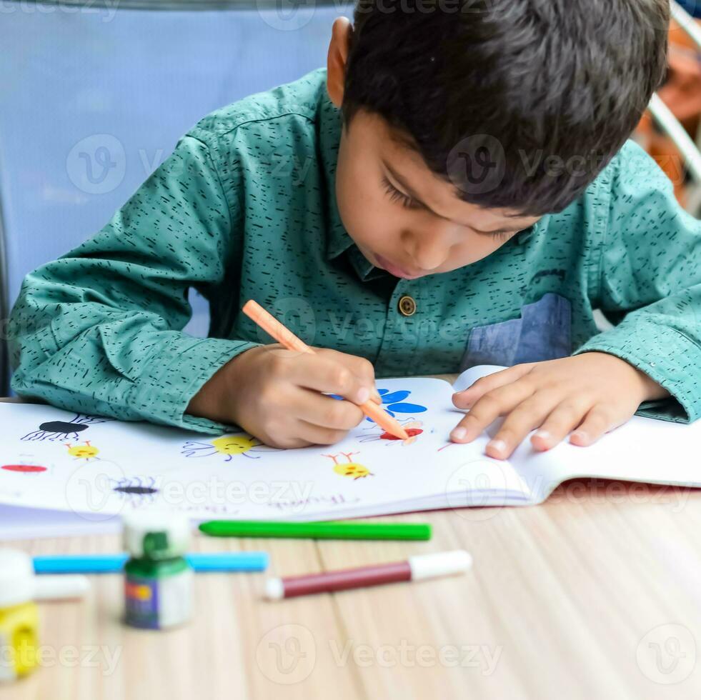 Smart Indian little boy perform thumb painting with different colourful water colour kit during the summer vacations, Cute Indian Kid doing colourful thumb painting drawing on wooden table photo