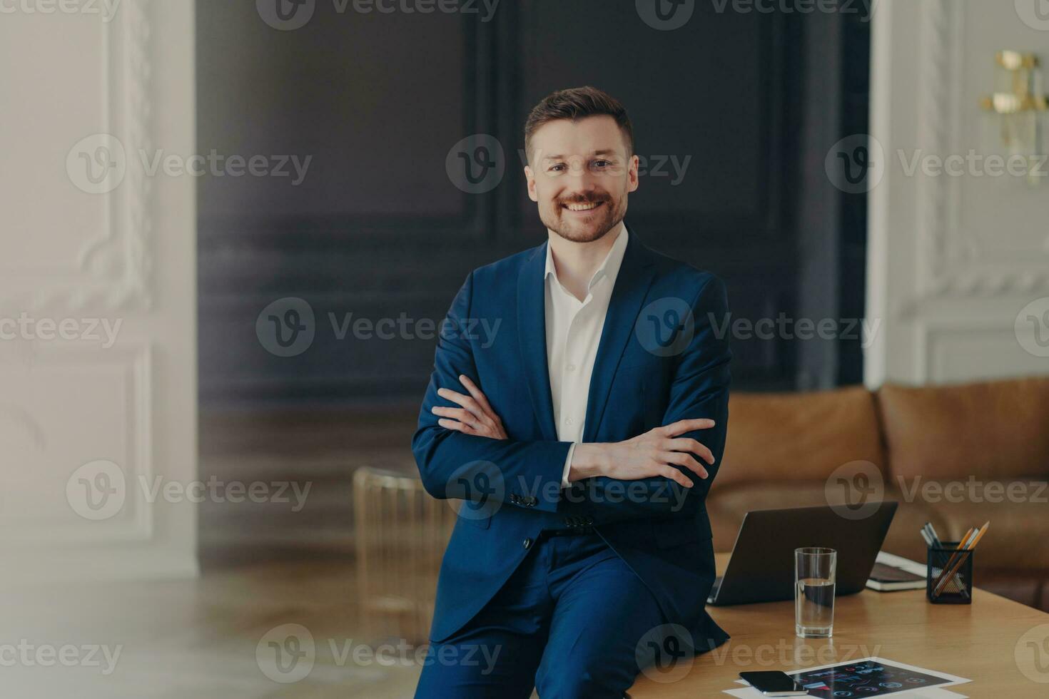 Satisfied bearded businessman in formal attire, stands confidently in office, arms folded, smiling pleasantly, content with work results. photo