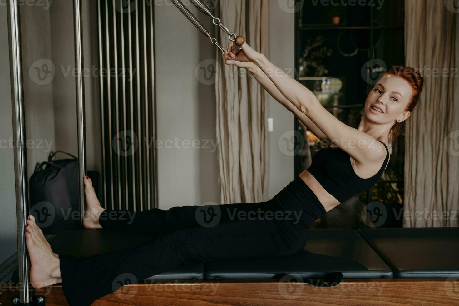 Redhead woman does arm stretching on Pilates reformer in studio. Smiling, enhancing muscle strength. Embracing healthy lifestyle and fitness. photo