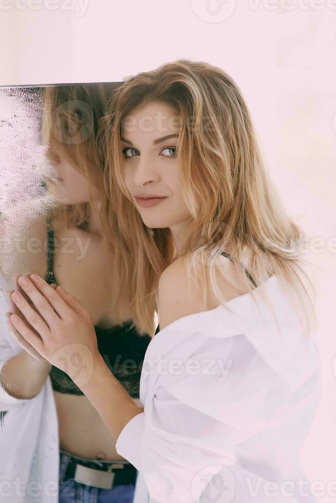 Beautiful caucasian blonde young woman in white shirt standing by the mirror. Health care, beauty, mental issues photo