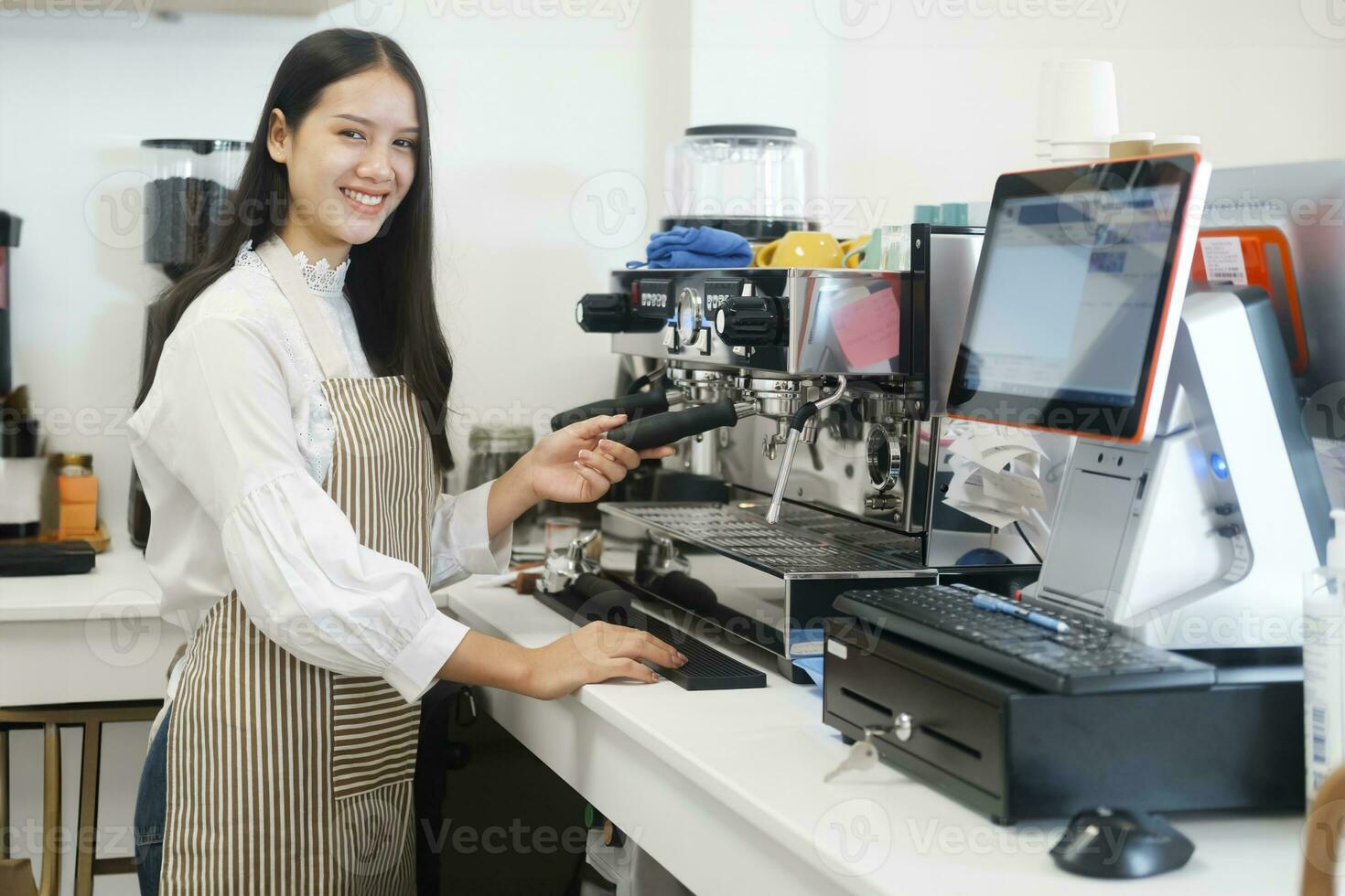 Young cheerful barista wearing apron while preparing coffee at an automatic machine in a modern coffee shop. photo