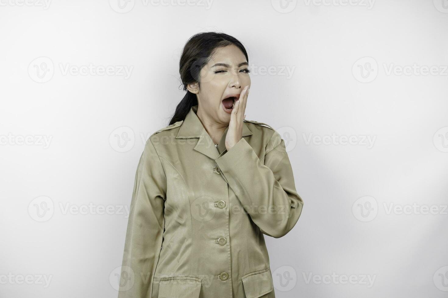 Portrait of sleepy Asian woman wearing a brown uniform and covering her mouth with a hand while yawning. Isolated image on white background photo