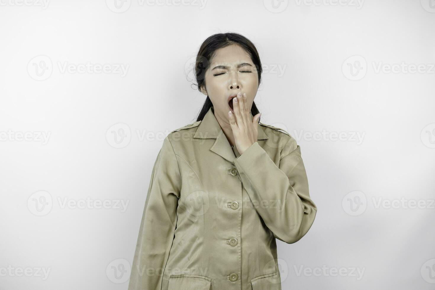 Portrait of sleepy Asian woman wearing a brown uniform and covering her mouth with a hand while yawning. Isolated image on white background photo