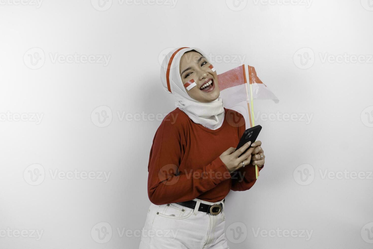 un sonriente asiático musulmán mujer vistiendo rojo parte superior y blanco hiyab, participación su teléfono y bandera, aislado por blanco antecedentes. de indonesia independencia día concepto foto