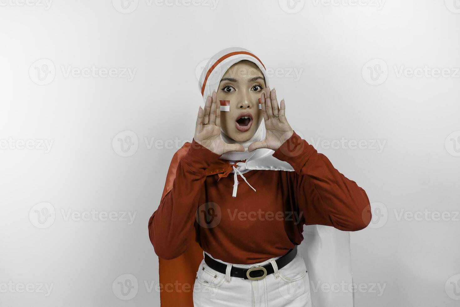 Young beautiful muslim woman wearing a red top and white hijab is holding Indonesia's flag and shouting and screaming loud with a hand on her mouth. Indonesia's independence day concept. photo