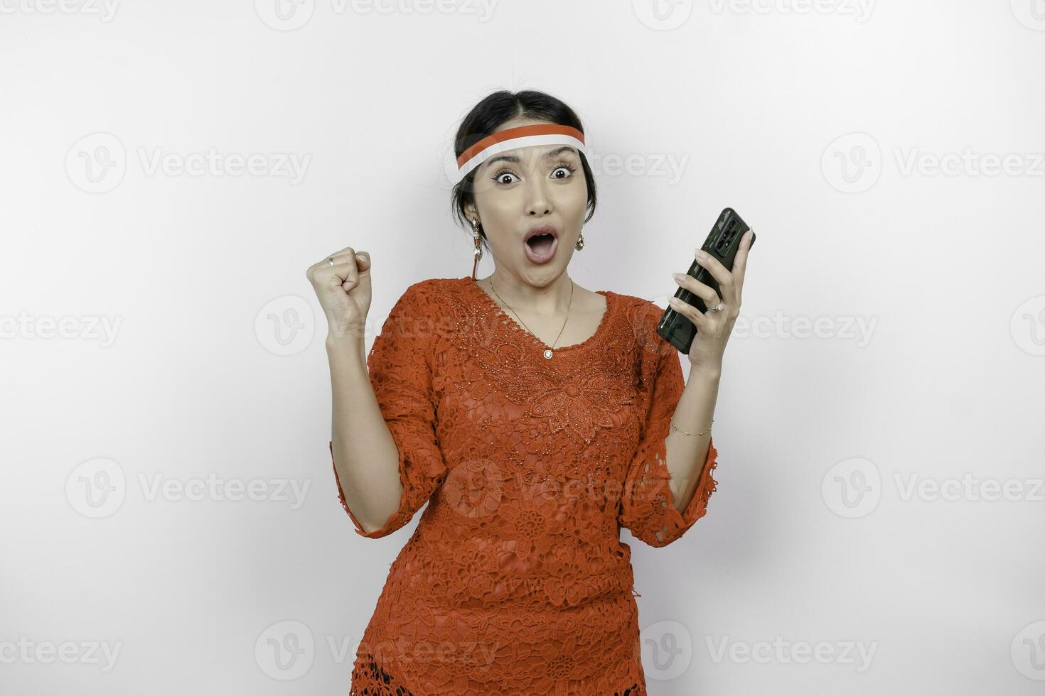 A young Asian woman with a happy successful expression while holding her phone and wearing red kebaya, flag headband isolated by white background. Indonesia's independence day concept. photo