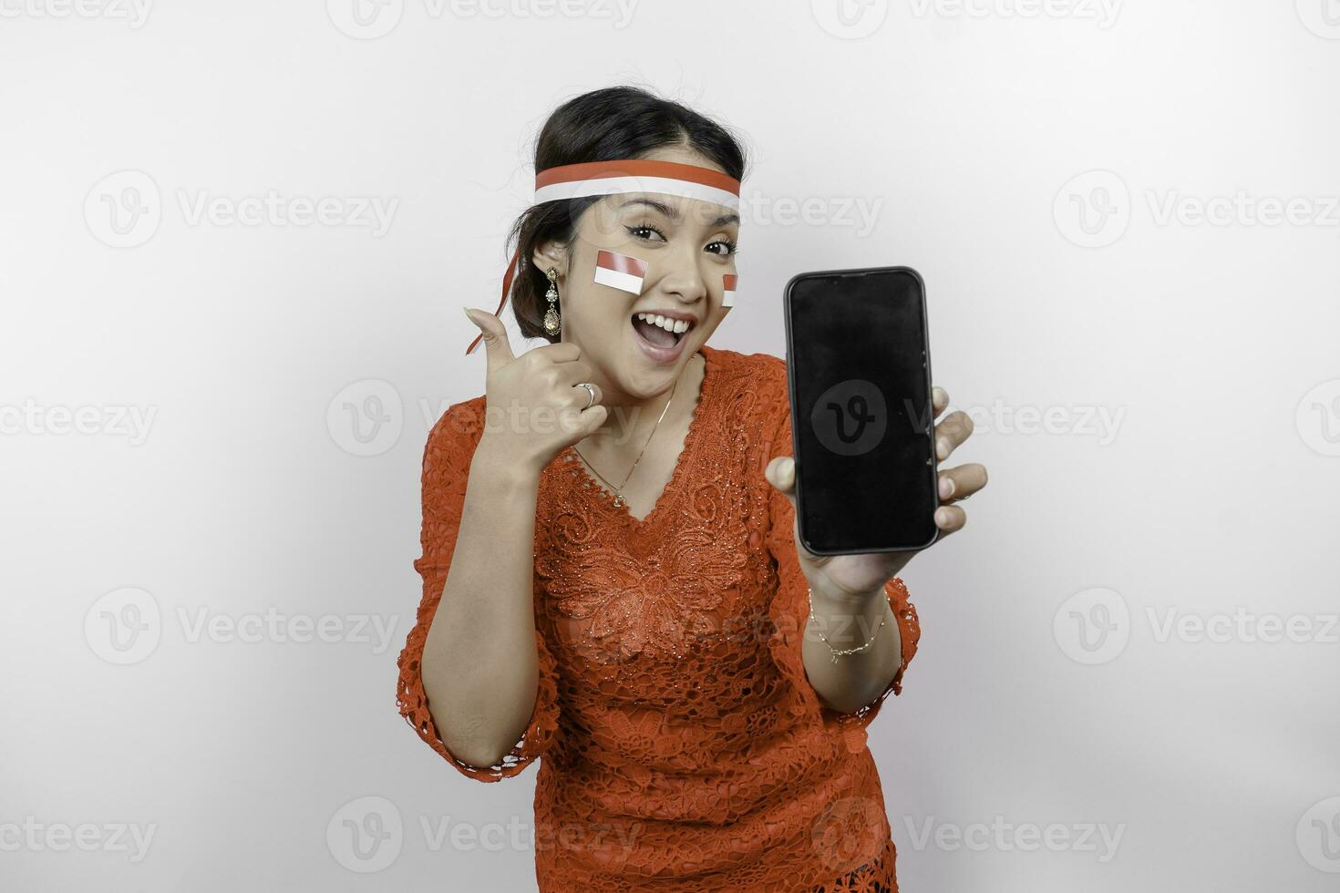 A cheerful Asian woman wearing red kebaya and headband, showing her phone while gesturing OK sign with her fingers, isolated by white background. Indonesia's independence day concept photo