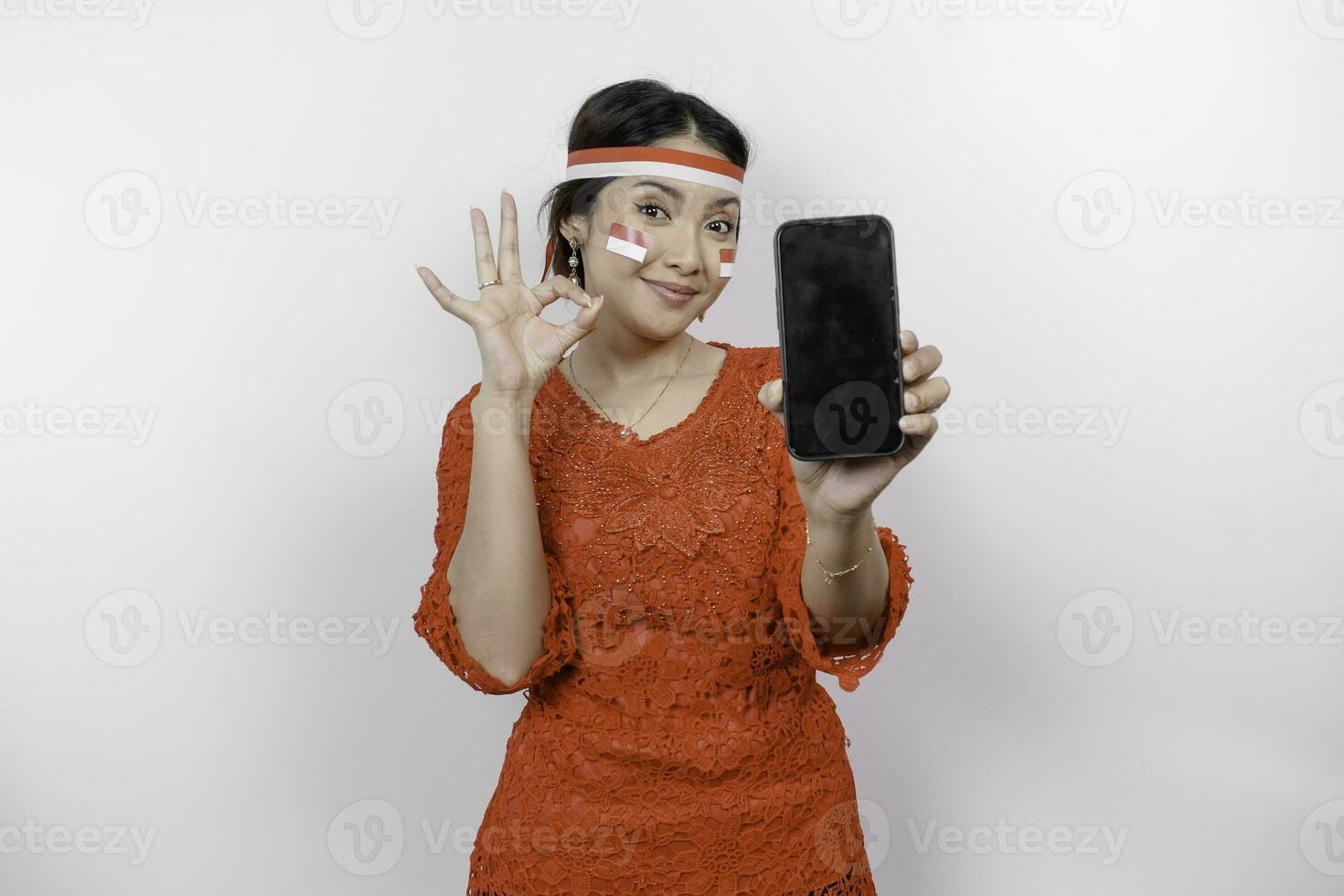 un alegre asiático mujer vistiendo rojo kebaya y venda, demostración su teléfono mientras gesticulando Okay firmar con su dedos, aislado por blanco antecedentes. de indonesia independencia día concepto foto