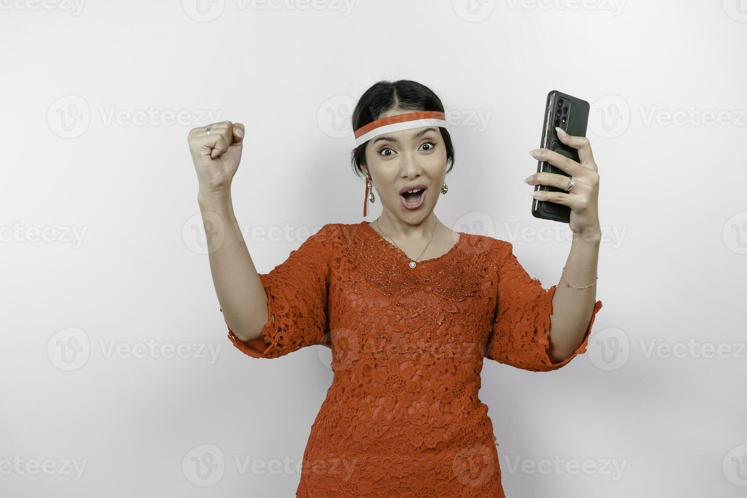 A young Asian woman with a happy successful expression while holding her phone and wearing red kebaya, flag headband isolated by white background. Indonesia's independence day concept. photo