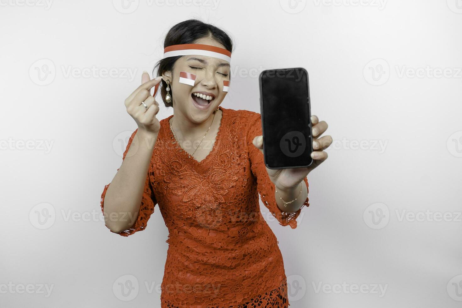 A happy Asian woman wearing red kebaya and headband, showing her phone, and gesturing love sign by her fingers showing tender feeling, isolated by white background. Indonesia's independence day photo