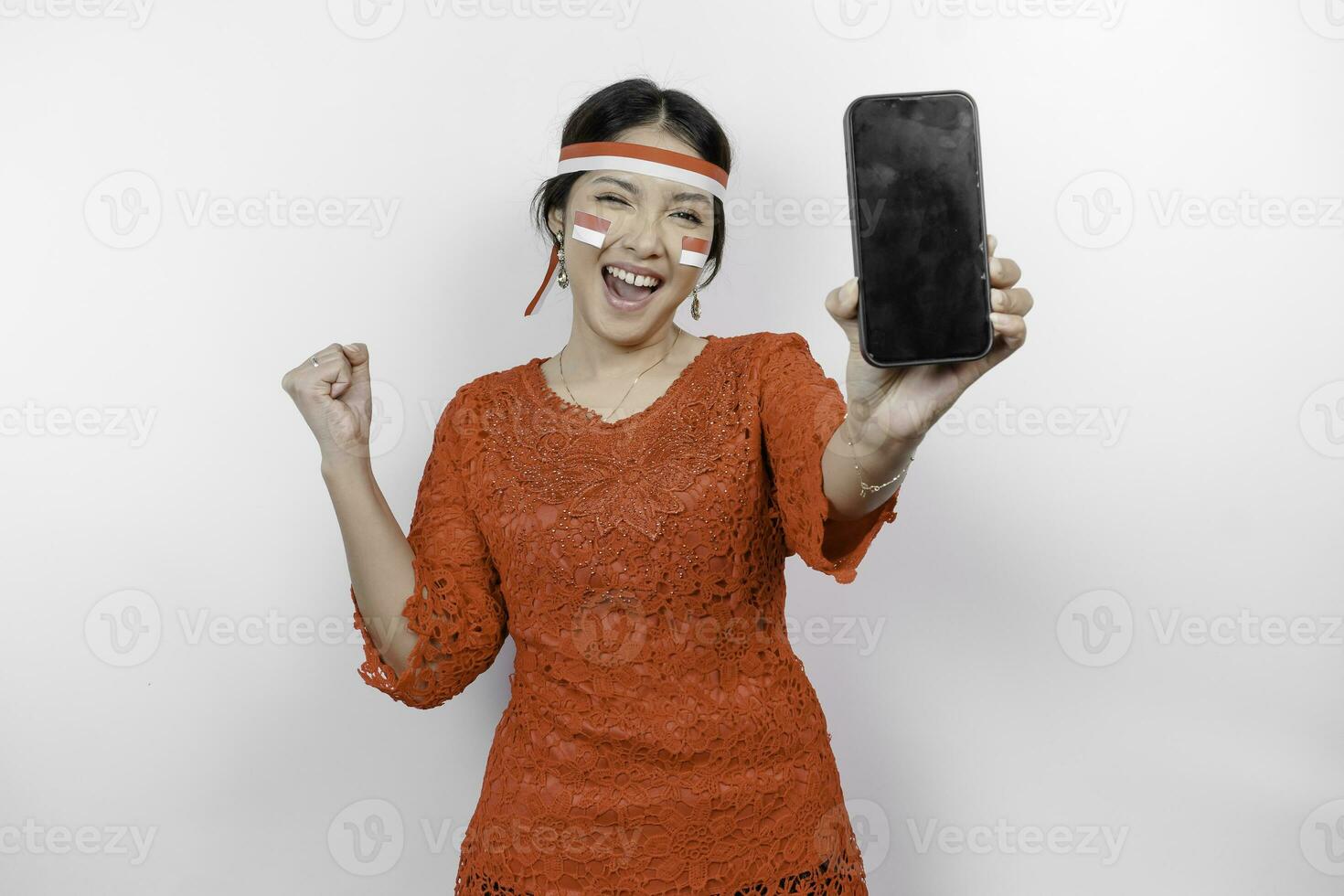 A young Asian woman with a happy successful expression while holding her phone and wearing red kebaya, flag headband isolated by white background. Indonesia's independence day concept. photo