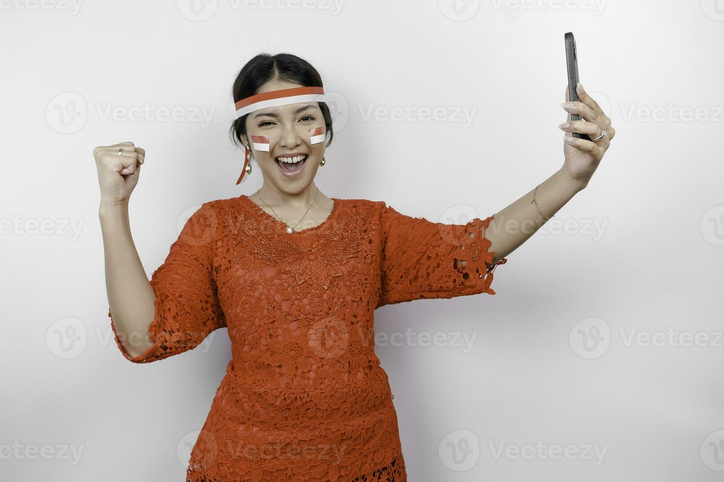 A young Asian woman with a happy successful expression while holding her phone and wearing red kebaya, flag headband isolated by white background. Indonesia's independence day concept. photo