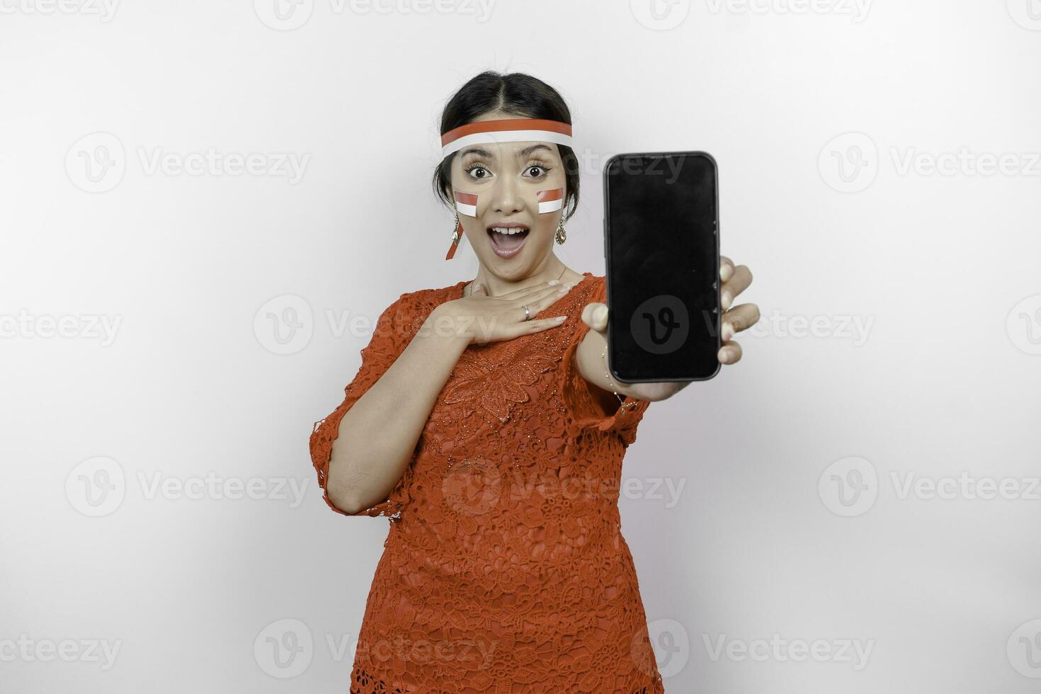 A portrait of a shocked Asian woman wearing red kebaya and headband, showing her phone while her mouth wide open, isolated by white background. Indonesia's independence day concept photo