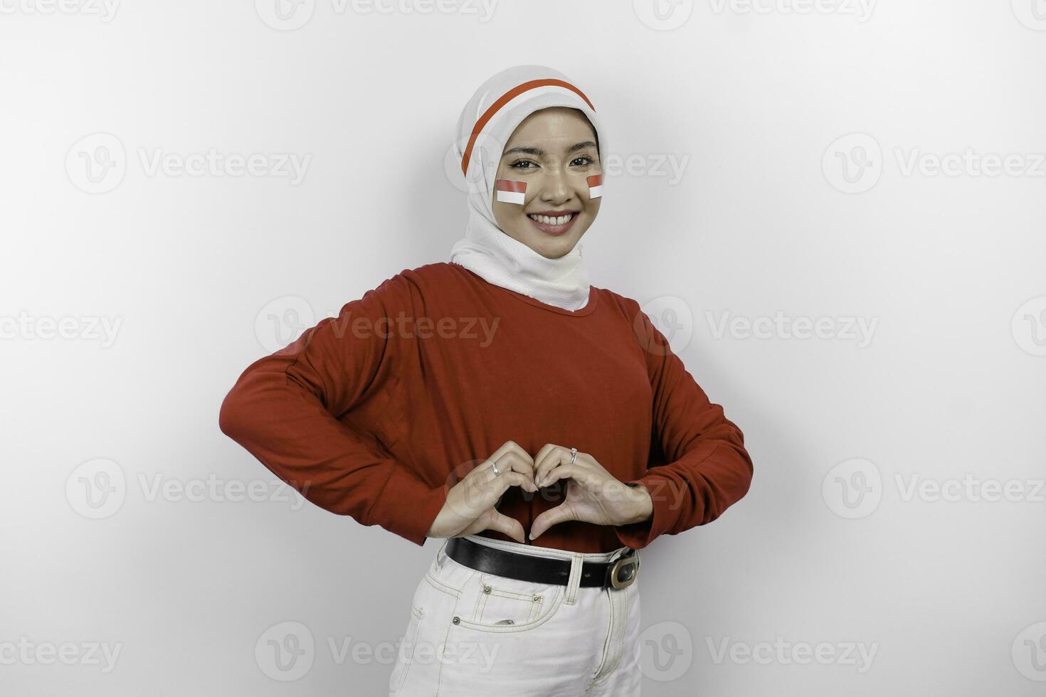 A young Asian woman wearing a red top and white hijab feels nationalism, shapes heart gesture expresses her love for Indonesia. Indonesia's independence day concept. photo