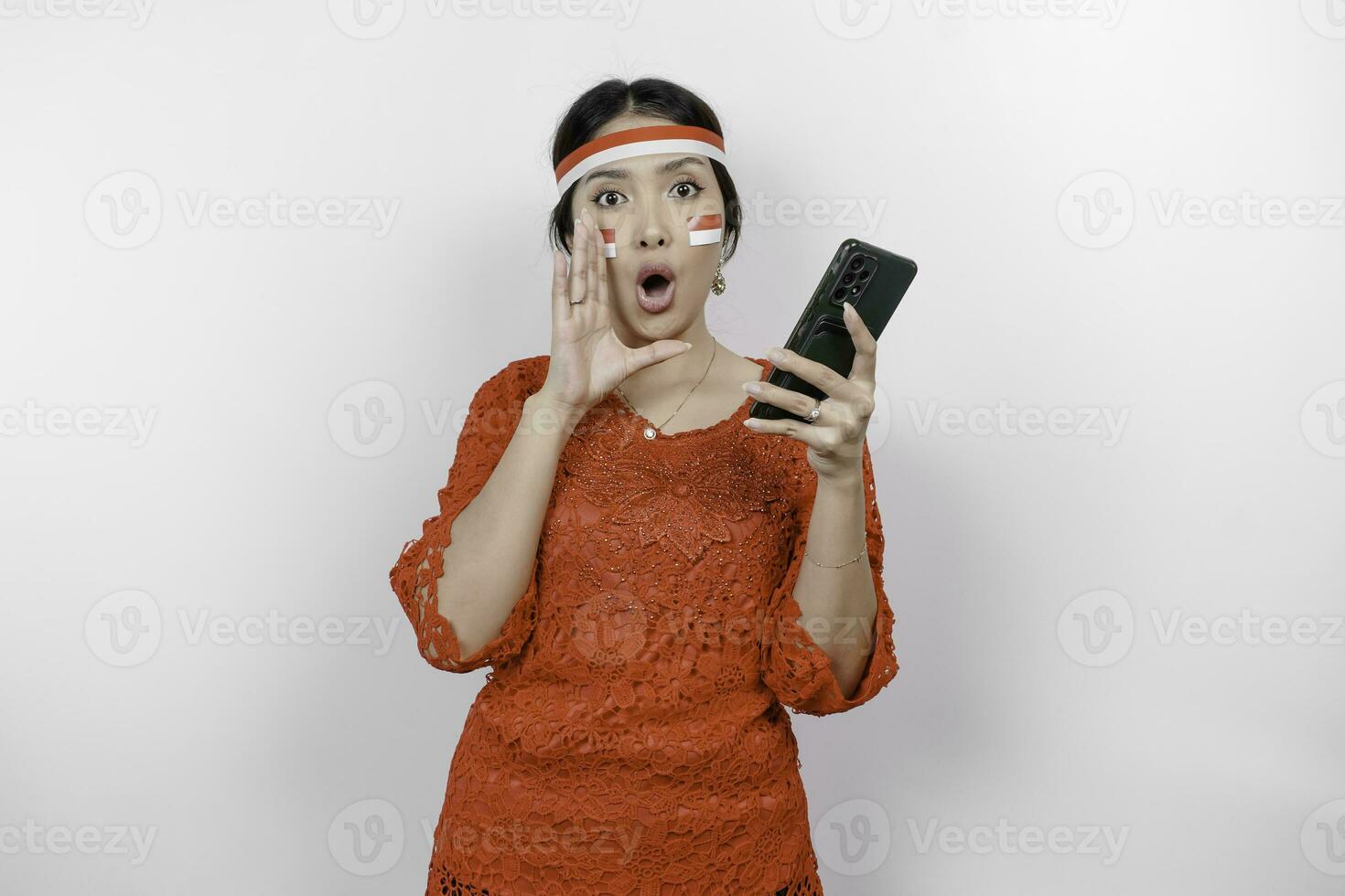 Young beautiful woman wearing a red kebaya is holding her phone while shouting and screaming loud with a hand on her mouth. Indonesia's independence day concept. photo