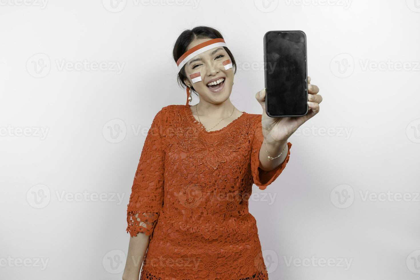 A portrait of a smiling Asian woman wearing red kebaya and headband and showing her phone, isolated by white background. Indonesia's independence day concept photo