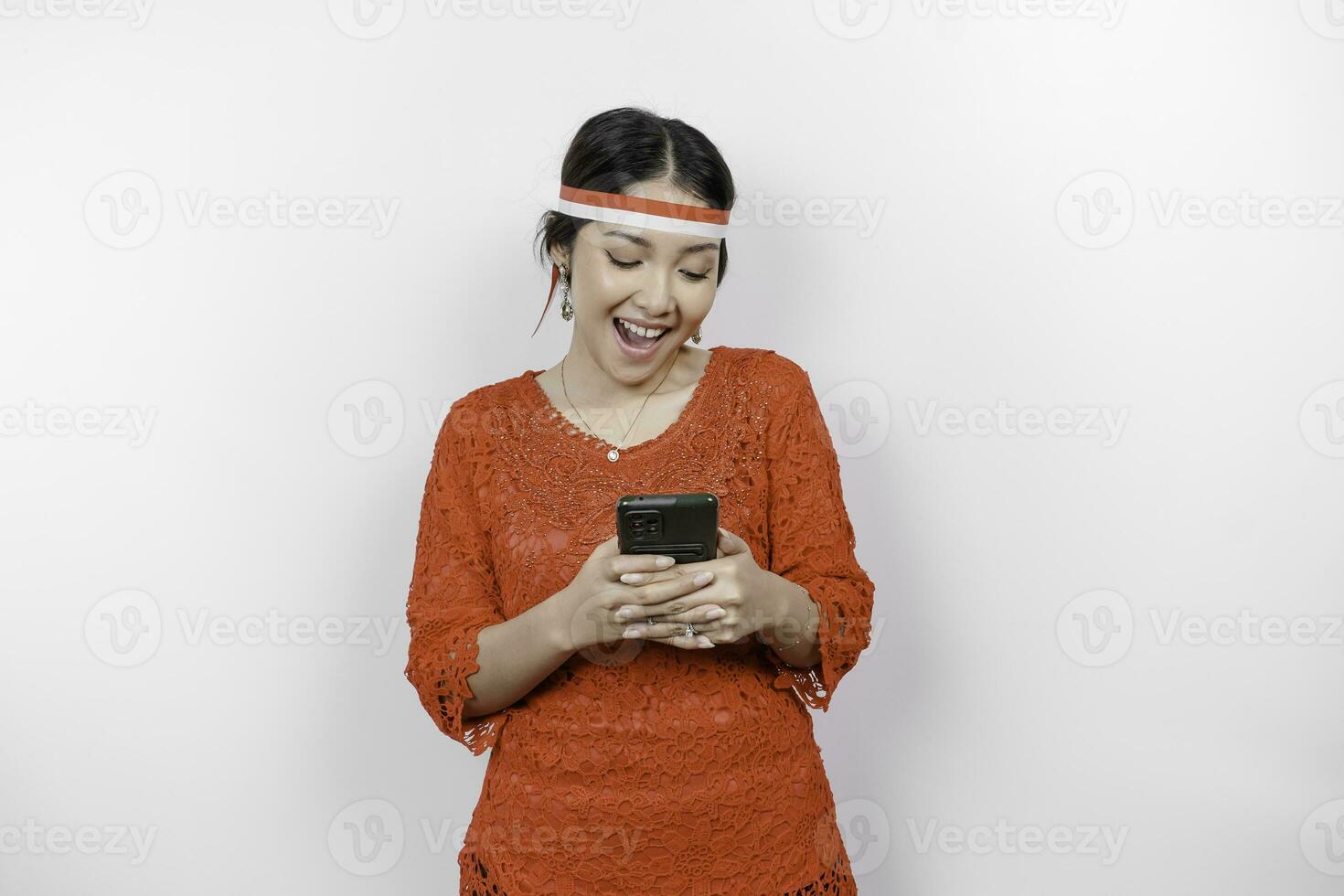 A portrait of a smiling Asian woman wearing red kebaya and headband and holding her phone, isolated by white background. Indonesia's independence day concept photo