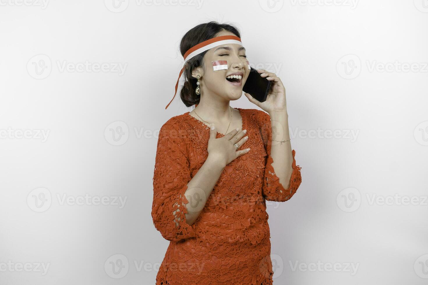 Happy mindful thankful young Asian woman wearing red kebaya and holding phone and hand on chest smiling isolated on white background feeling no stress, photo