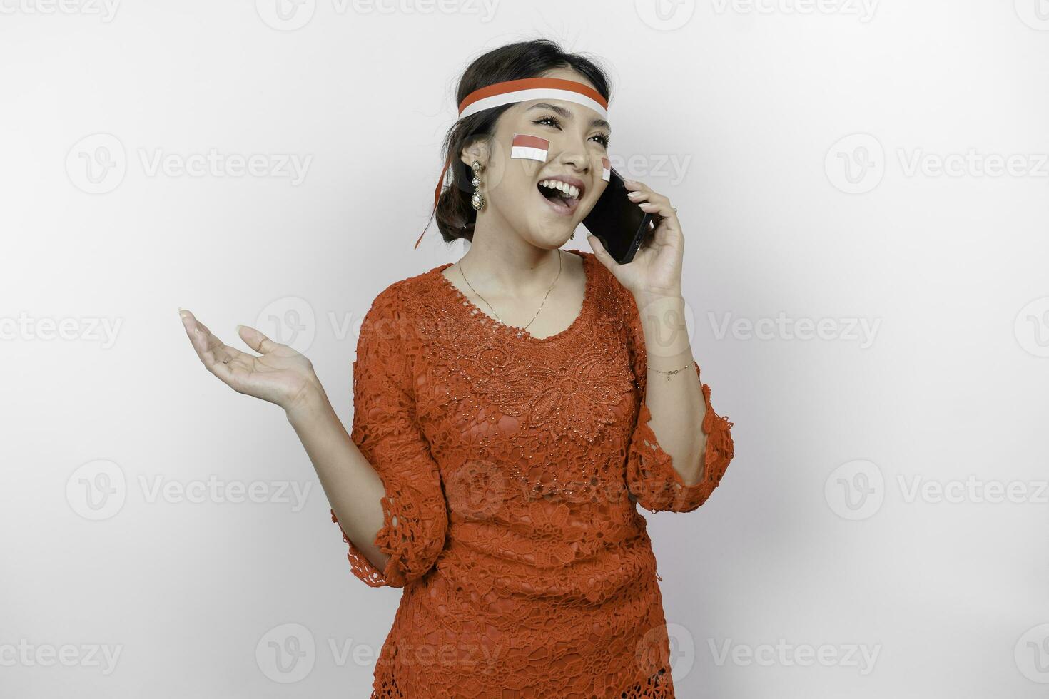 A happy Asian woman wearing red kebaya and headband, holding her phone, and pointing copy space beside her, isolated by white background. Indonesia's independence day photo