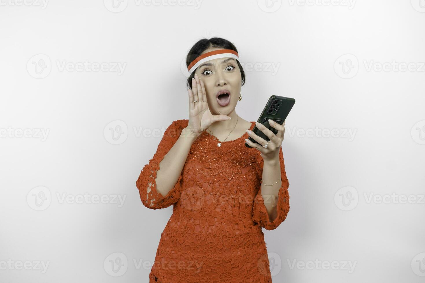 Young beautiful woman wearing a red kebaya is holding her phone while shouting and screaming loud with a hand on her mouth. Indonesia's independence day concept. photo