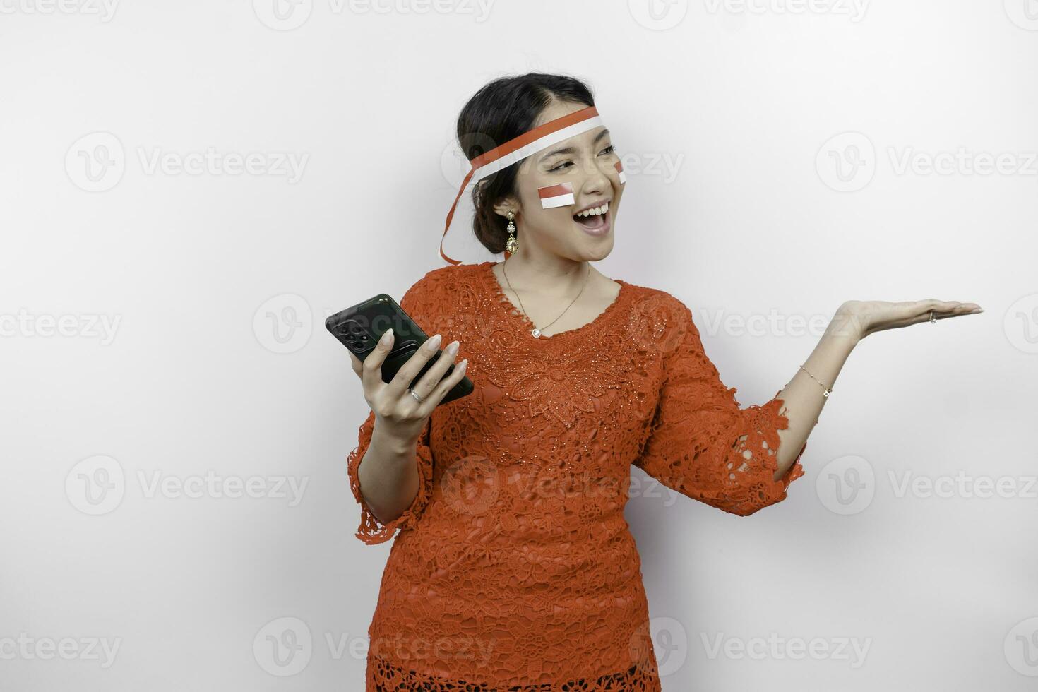 A happy Asian woman wearing red kebaya and headband, holding her phone, and pointing copy space beside her, isolated by white background. Indonesia's independence day photo