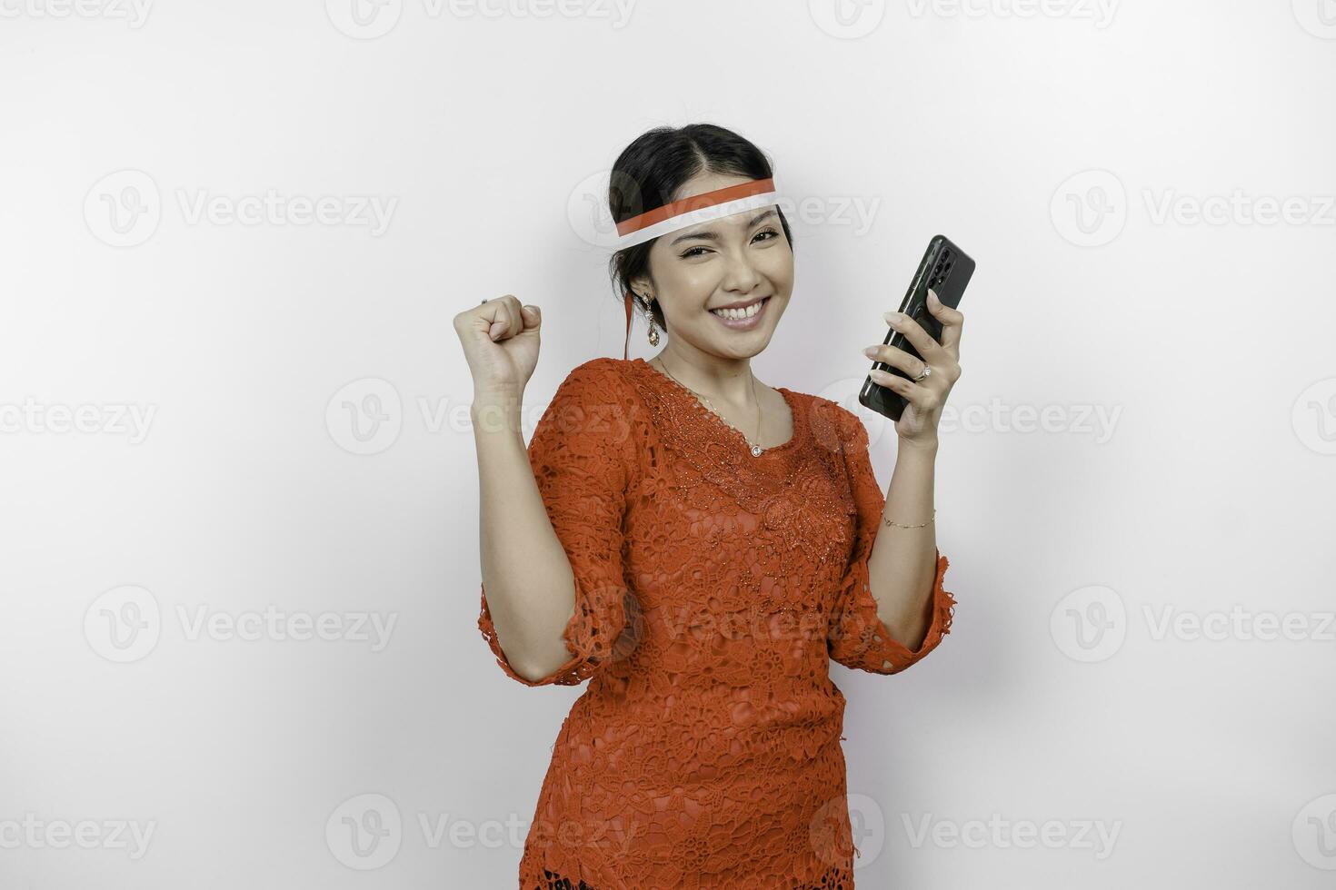 A young Asian woman with a happy successful expression while holding her phone and wearing red kebaya, flag headband isolated by white background. Indonesia's independence day concept. photo
