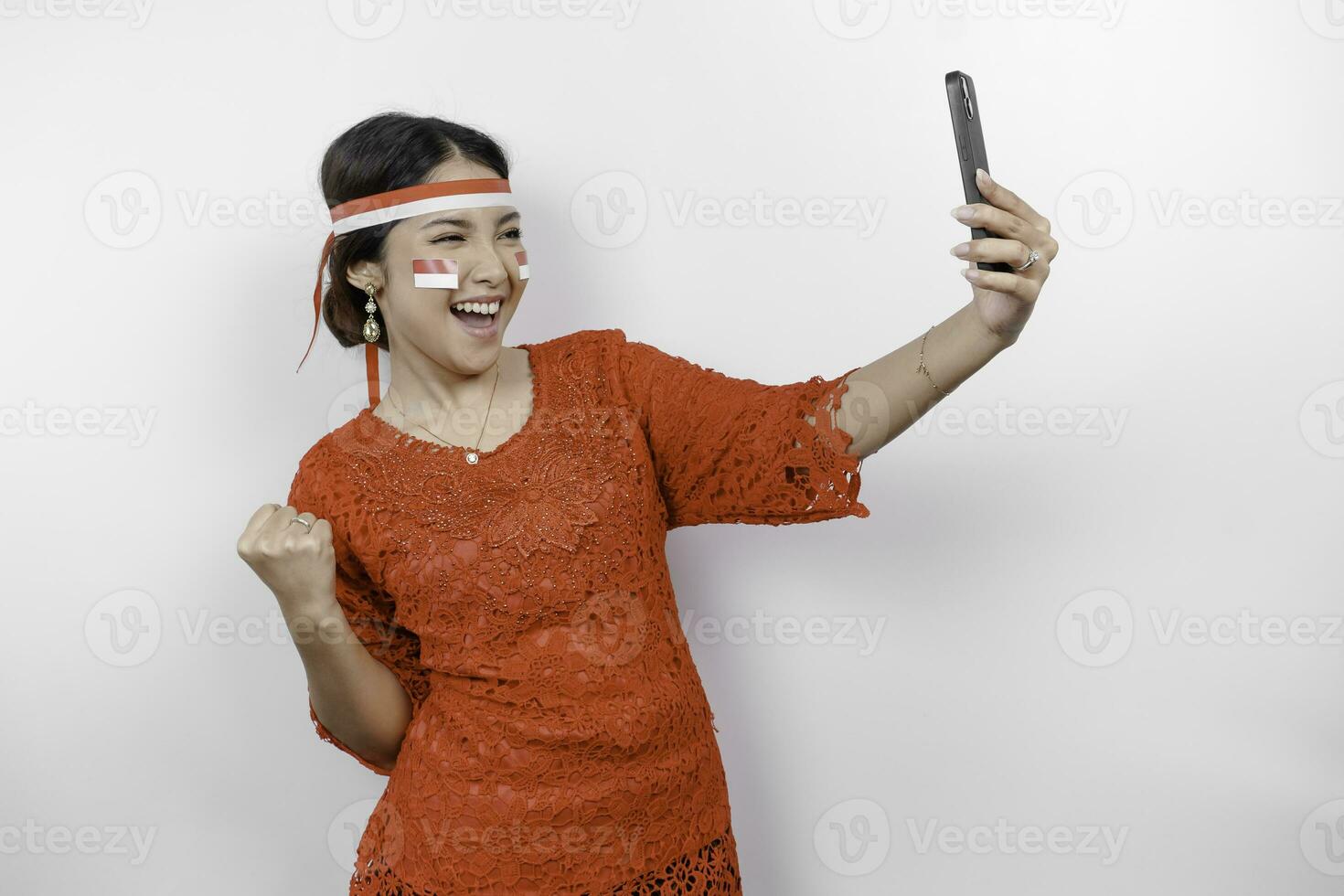 A young Asian woman with a happy successful expression while holding her phone and wearing red kebaya, flag headband isolated by white background. Indonesia's independence day concept. photo