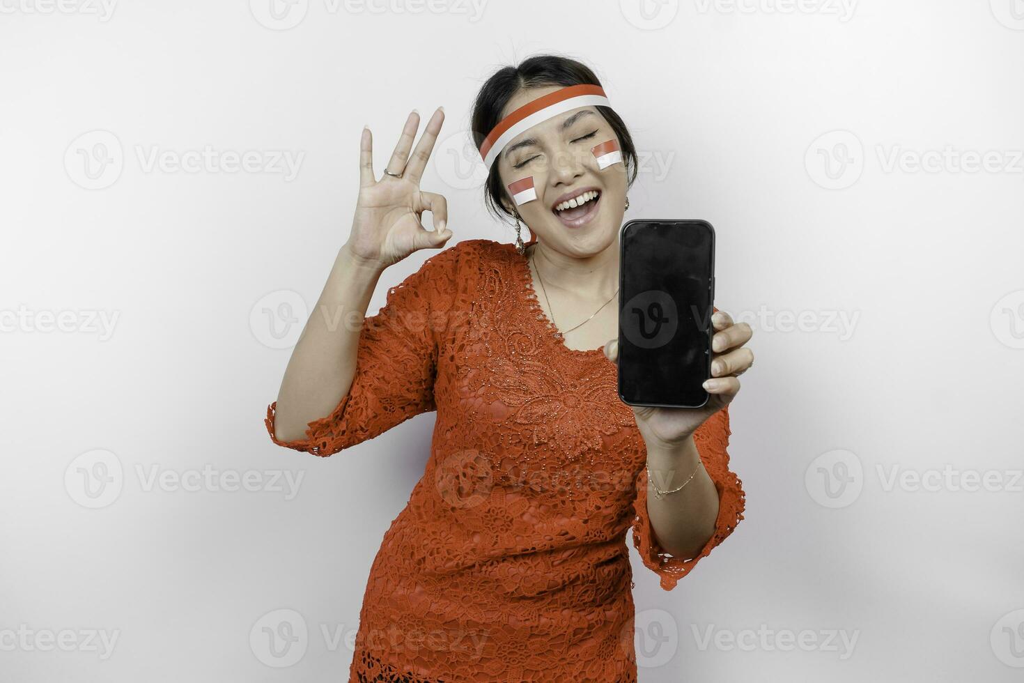 A cheerful Asian woman wearing red kebaya and headband, showing her phone while gesturing OK sign with her fingers, isolated by white background. Indonesia's independence day concept photo