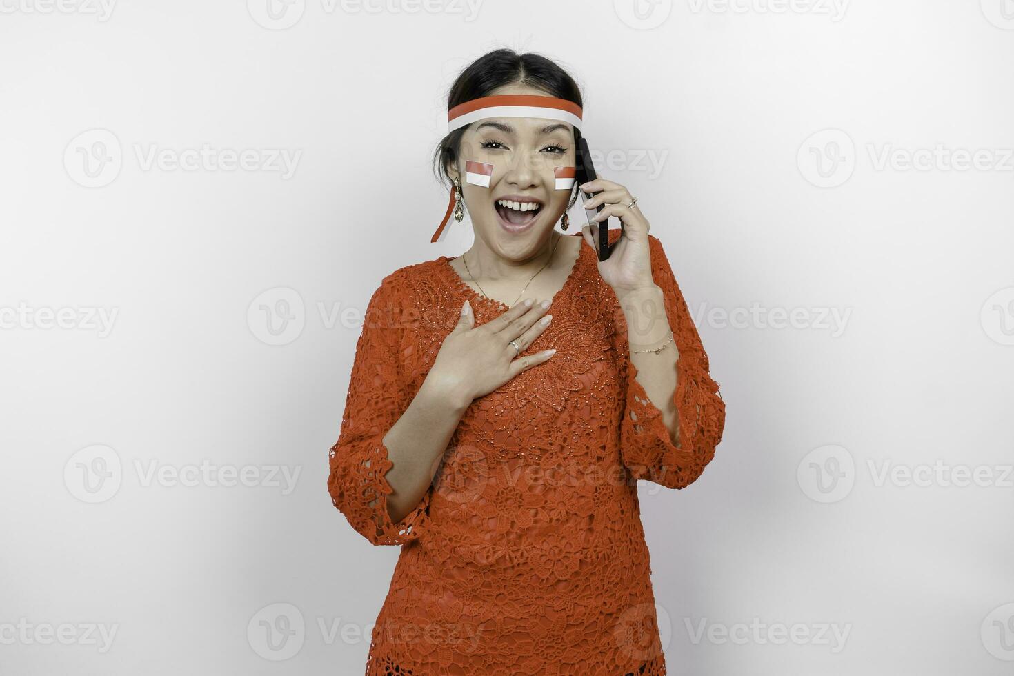 Happy mindful thankful young Asian woman wearing red kebaya and holding phone and hand on chest smiling isolated on white background feeling no stress, photo