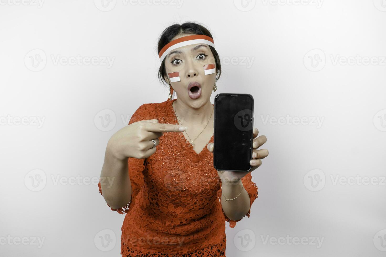 A portrait of a shocked Asian woman wearing red kebaya and headband, showing her phone while her mouth wide open, isolated by white background. Indonesia's independence day concept photo