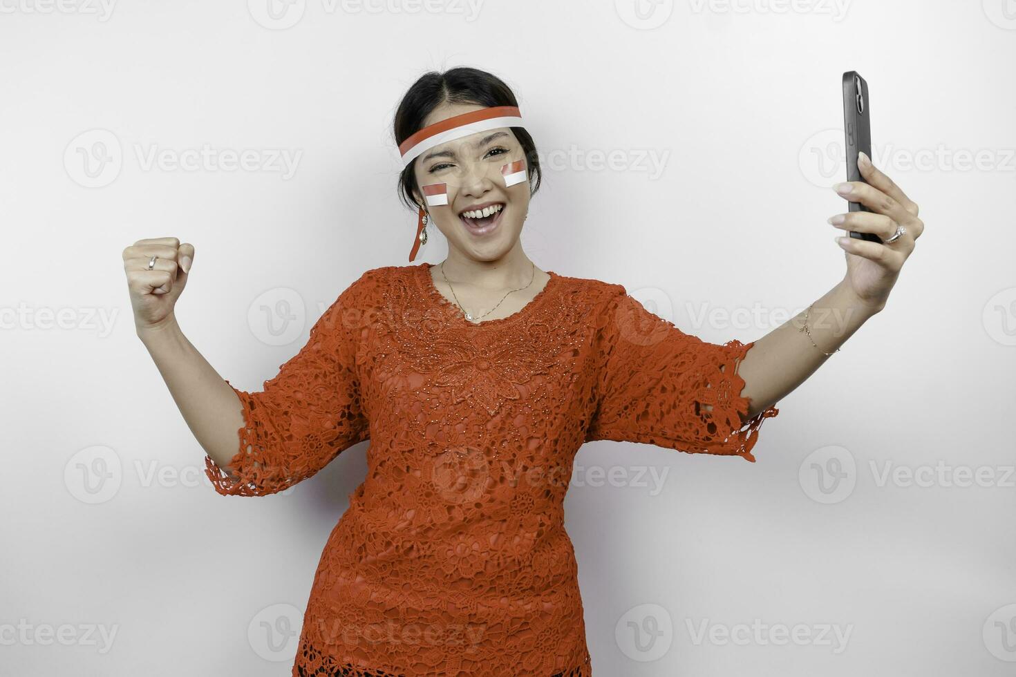 A young Asian woman with a happy successful expression while holding her phone and wearing red kebaya, flag headband isolated by white background. Indonesia's independence day concept. photo