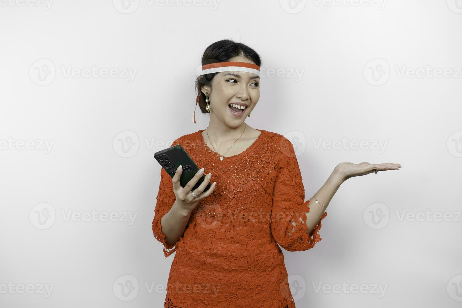 A happy Asian woman wearing red kebaya and headband, holding her phone, and pointing copy space beside her, isolated by white background. Indonesia's independence day photo