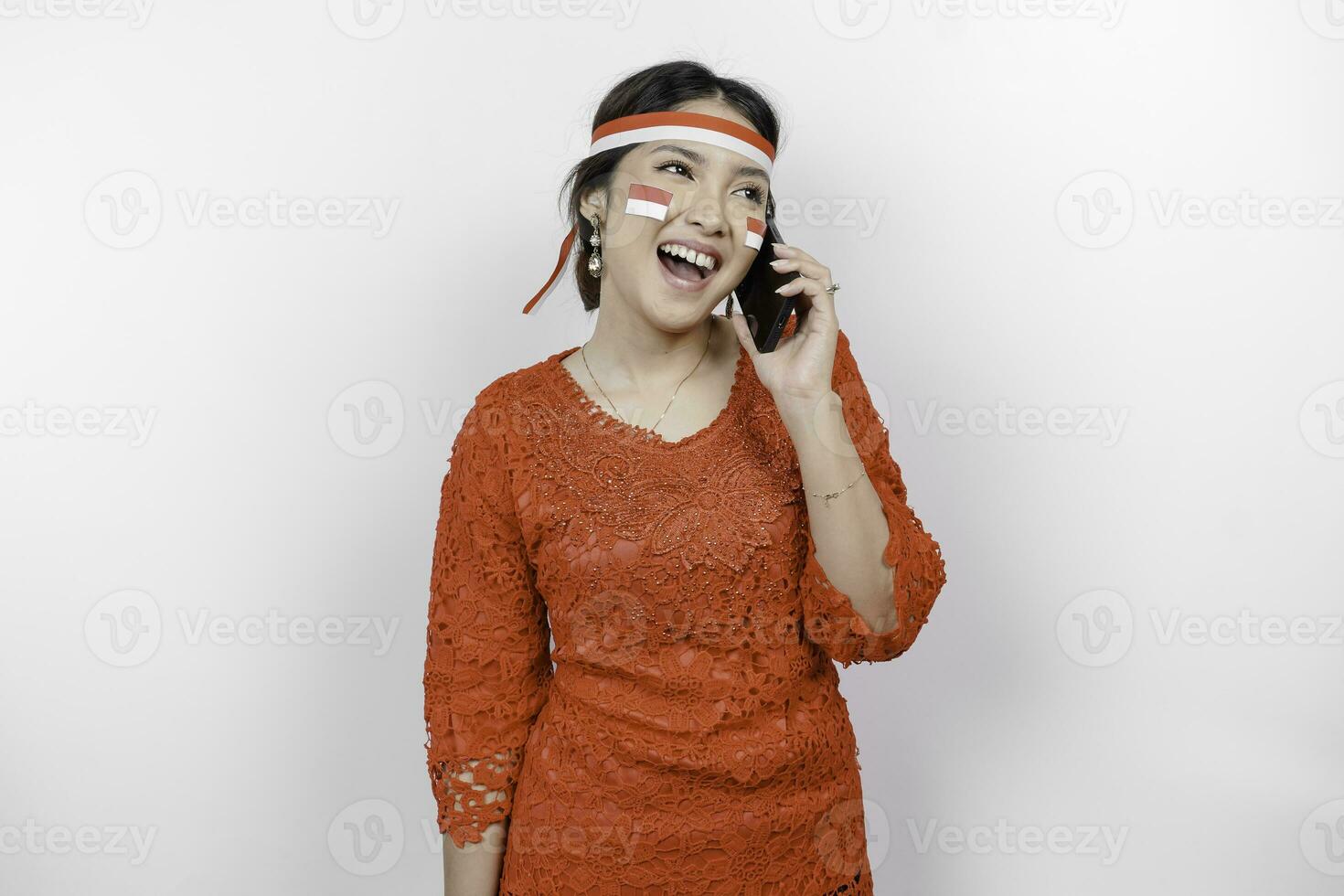 A portrait of a smiling Asian woman wearing red kebaya and headband and holding her phone, isolated by white background. Indonesia's independence day concept photo