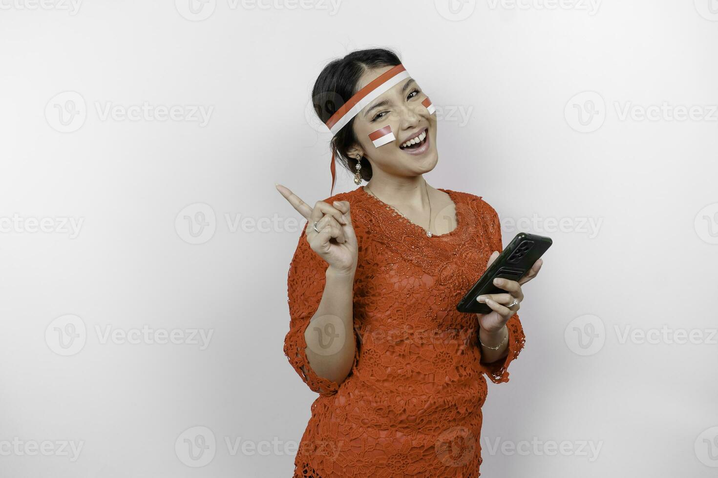 A happy Asian woman wearing red kebaya and headband, holding her phone, and pointing copy space on top of her, isolated by white background. Indonesia's independence day photo