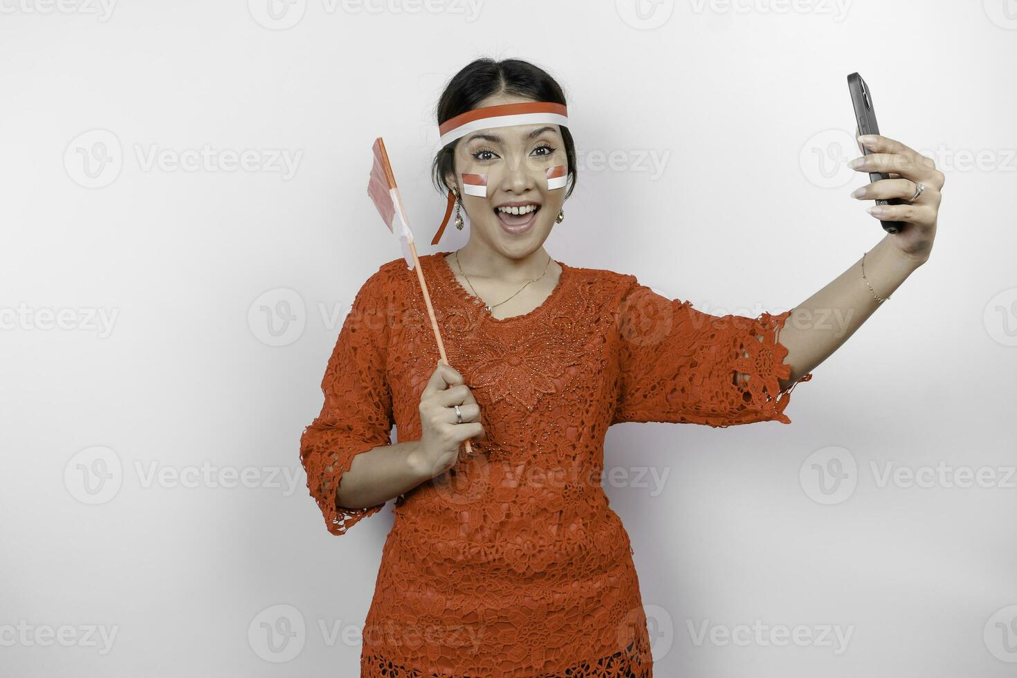 A happy Asian woman wearing red kebaya and headband, holding her phone and Indonesia's flag, isolated by white background. Indonesia's independence day photo