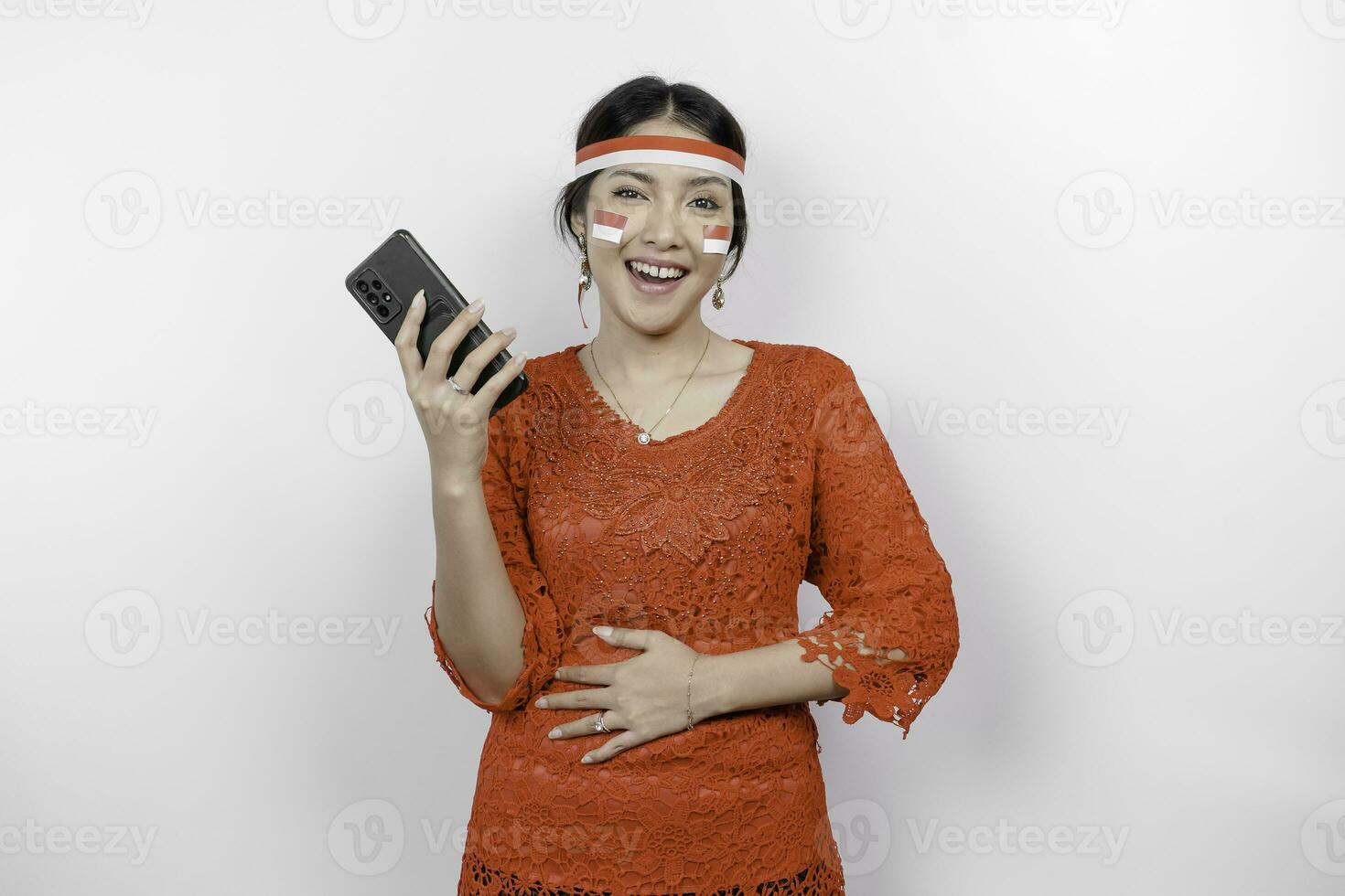 A thoughtful Asian woman wearing red kebaya and headband, holding her phone and stomach, thinking about what to eat or order, isolated by white background. Indonesia's independence day photo