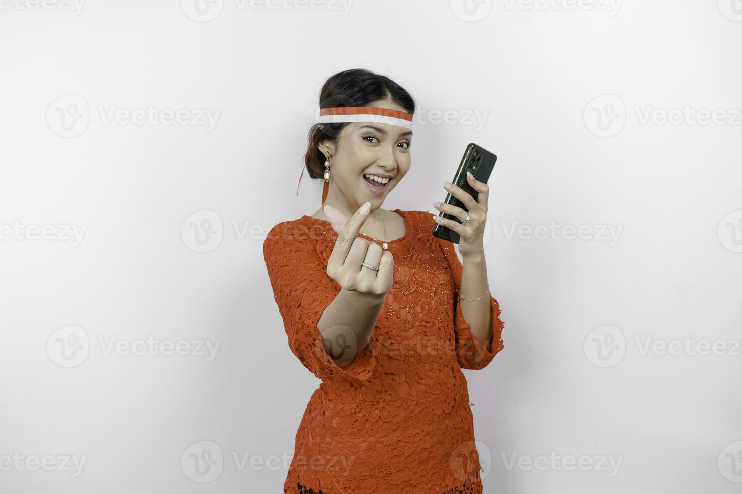 A happy Asian woman wearing red kebaya and headband, holding her phone, and gesturing love sign by her fingers showing tender feeling, isolated by white background. Indonesia's independence day photo