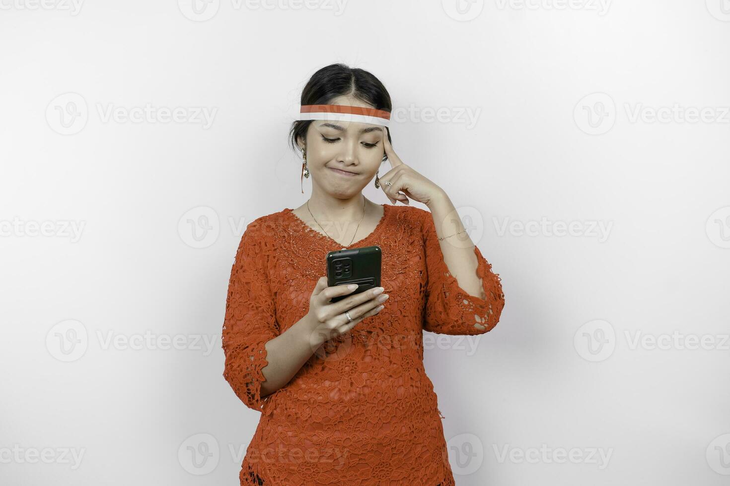 A smart young woman wearing headband and red kebaya is looking aside to an idea on copy space while holding her phone, isolated by white background. Indonesia's independence day. photo