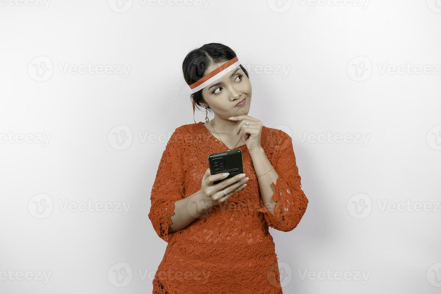 A smart young woman wearing headband and red kebaya is looking aside to an idea on copy space while holding her phone, isolated by white background. Indonesia's independence day. photo