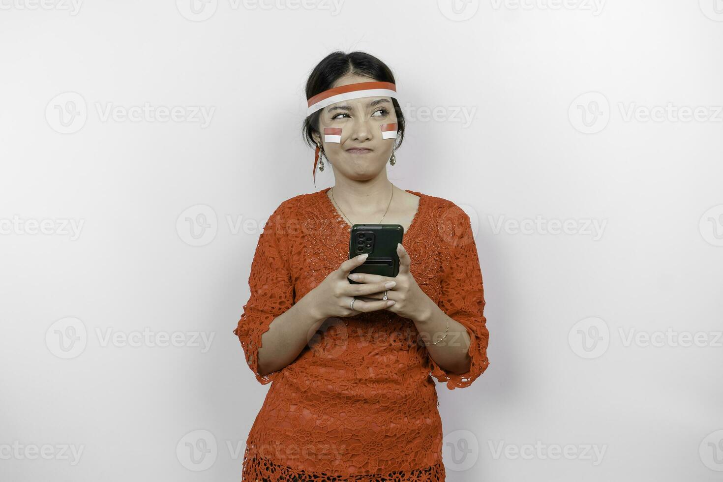 un inteligente joven mujer vistiendo venda y rojo kebaya es mirando aparte a un idea en Copiar espacio mientras participación su teléfono, aislado por blanco antecedentes. de indonesia independencia día. foto